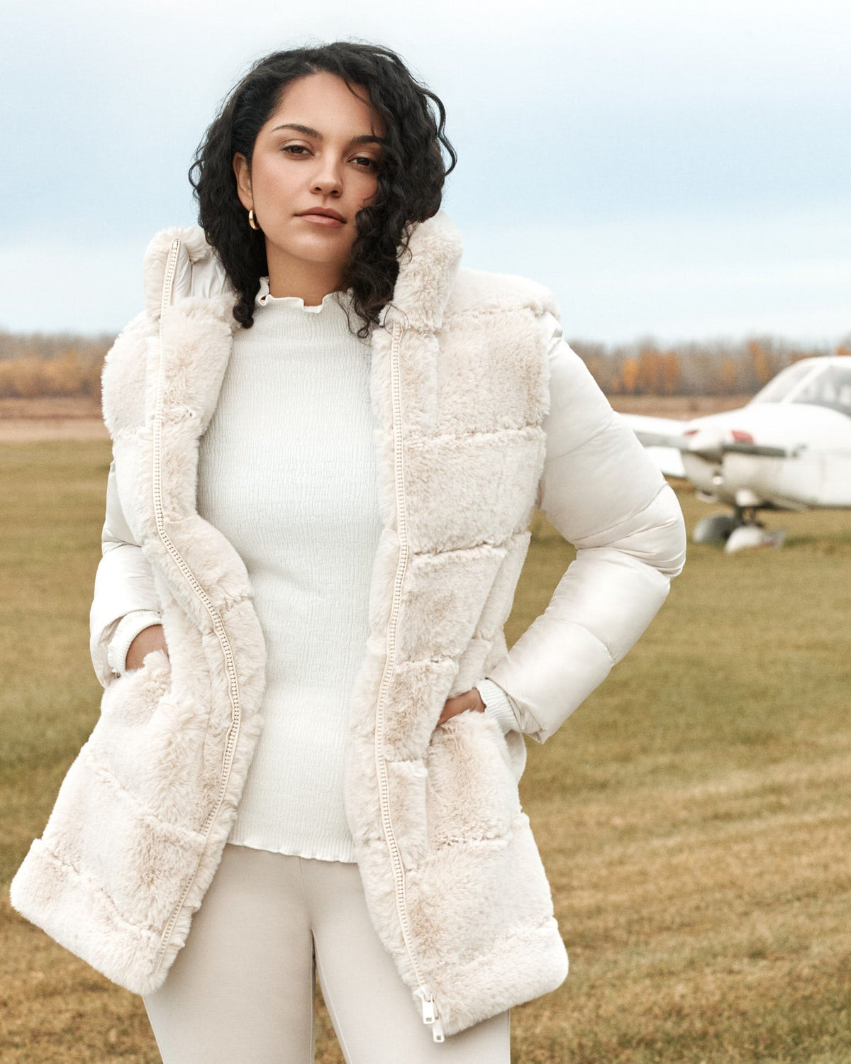 Woman in a white coat standing in front of a small airplane on a grassy field.